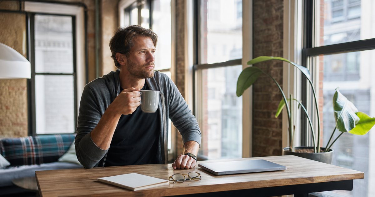 A writer at a standing desk in a sunlit loft apartment, pausing thoughtfully with a mug in hand, a notebook and reading glasses beside a closed laptop