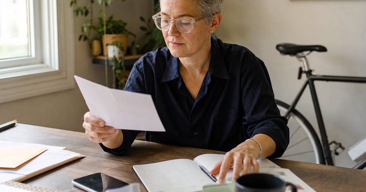 A person in a small home office reading a letter at a desk by the window, one hand resting on a paper planner, a bicycle leaning against the wall behind them